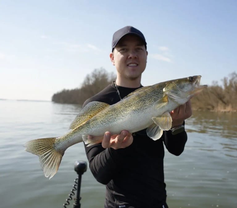 Een gast met een snoekbaars, met de kale voorjaarsbomen op de oever erachter