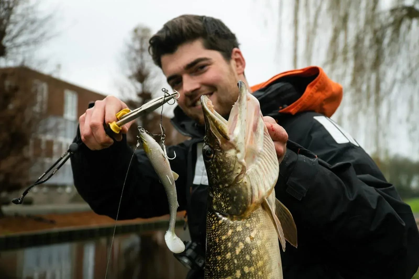 Een visser houdt een snoek rechtop bij de kieuwdeksel en maakt hem los met een onthaaktang, met het witte shad nog aan de lijn