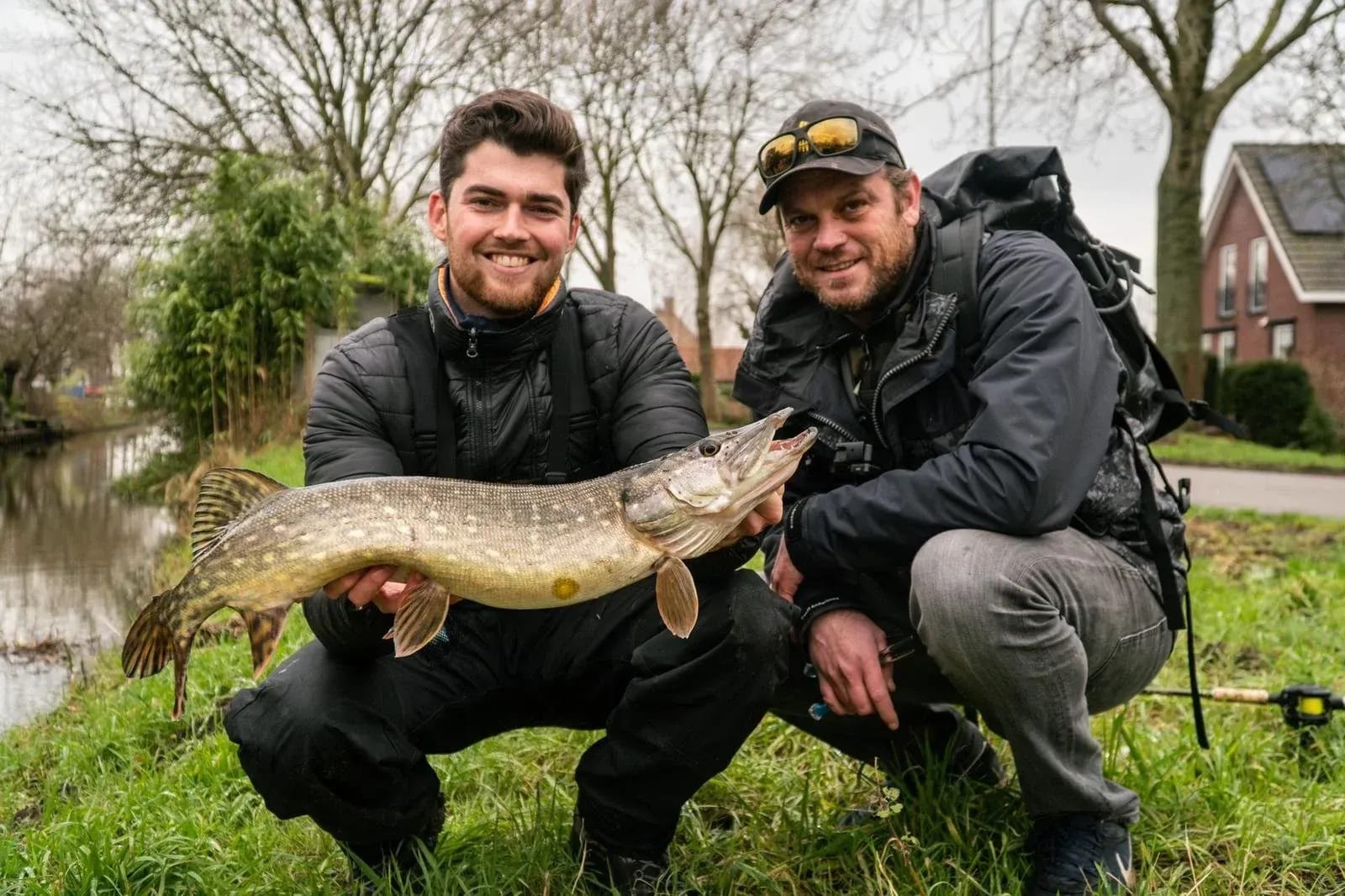 Twee vissers zitten gehurkt in het gras langs een poldersloot en houden samen een snoek vast, met huizen en kale bomen erachter