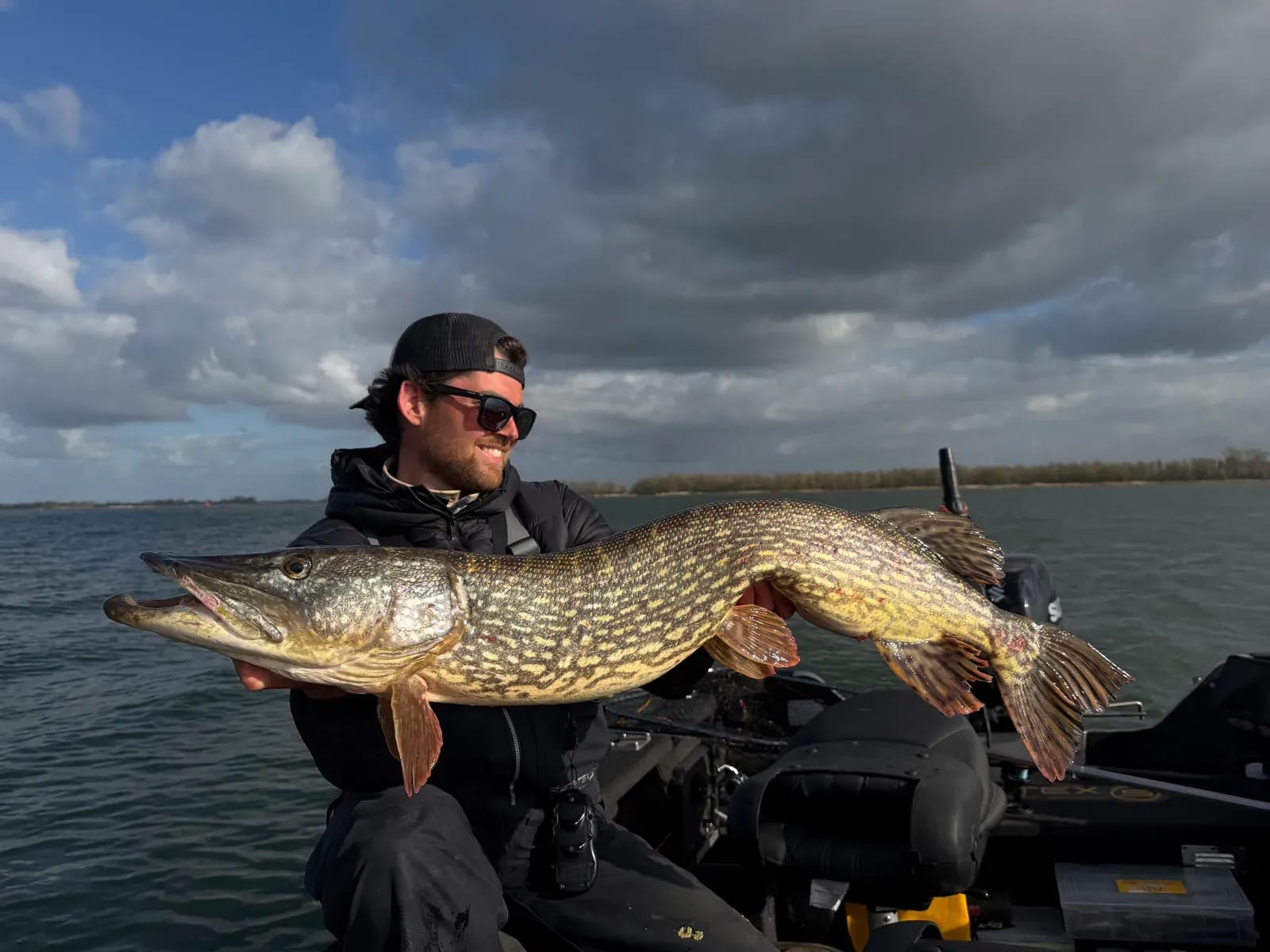 Luc met een kapitale snoek op open water, met doorbrekende zon achter de wolken