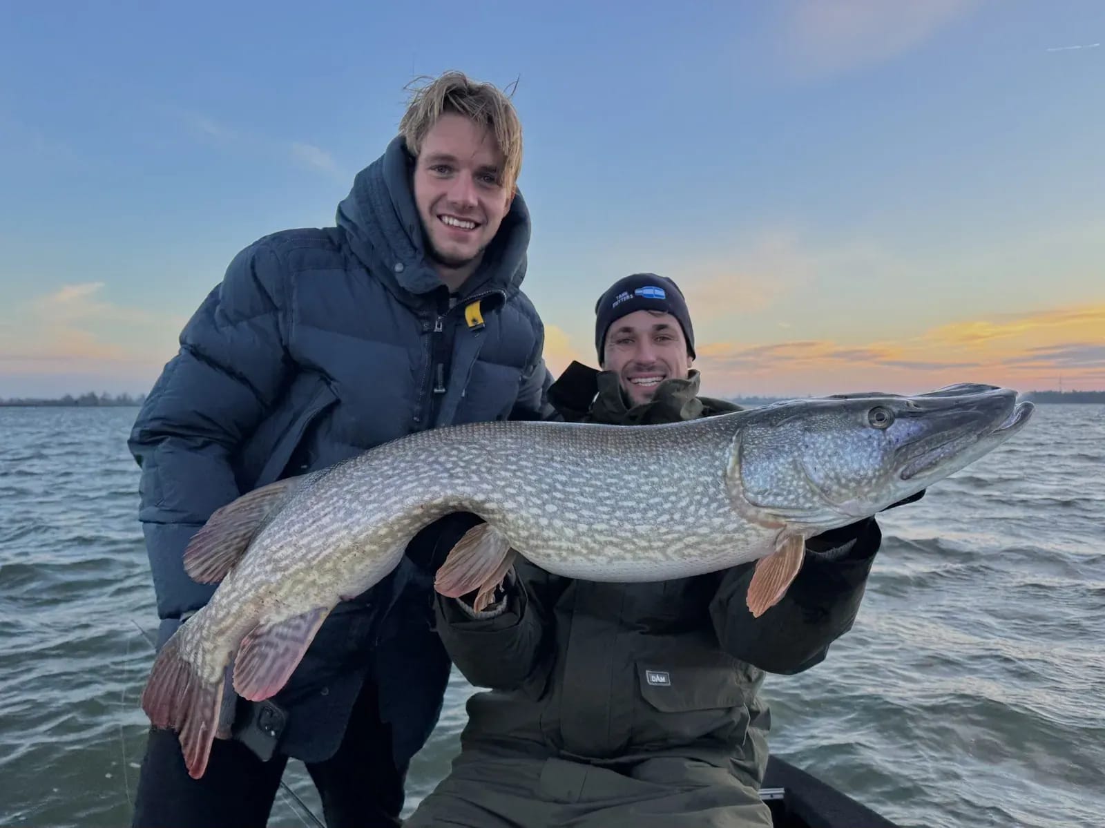 Twee vissers in winterkleding houden de snoek van 117 centimeter vast tegen een oranje avondlucht