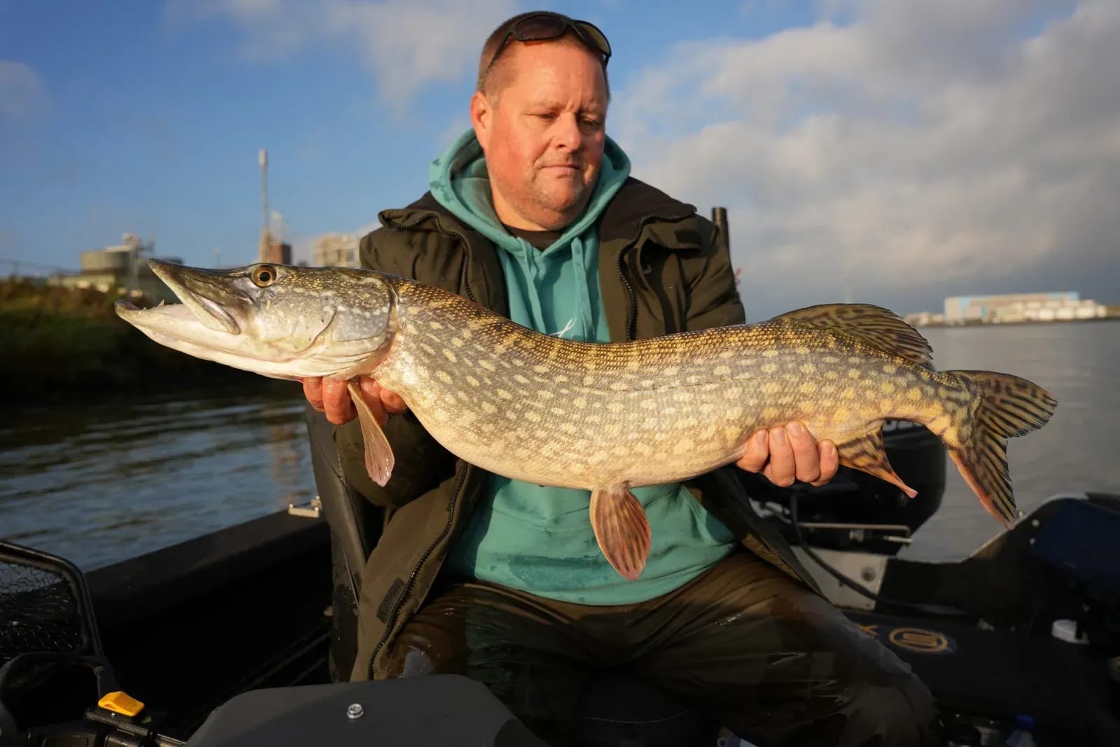 Dennis met een snoek van bijna een meter, met de industrie langs de rivier op de achtergrond